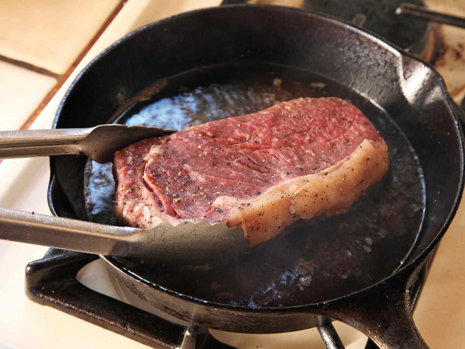 Steak being cooked and turned with tongs on a cast iron skillet.