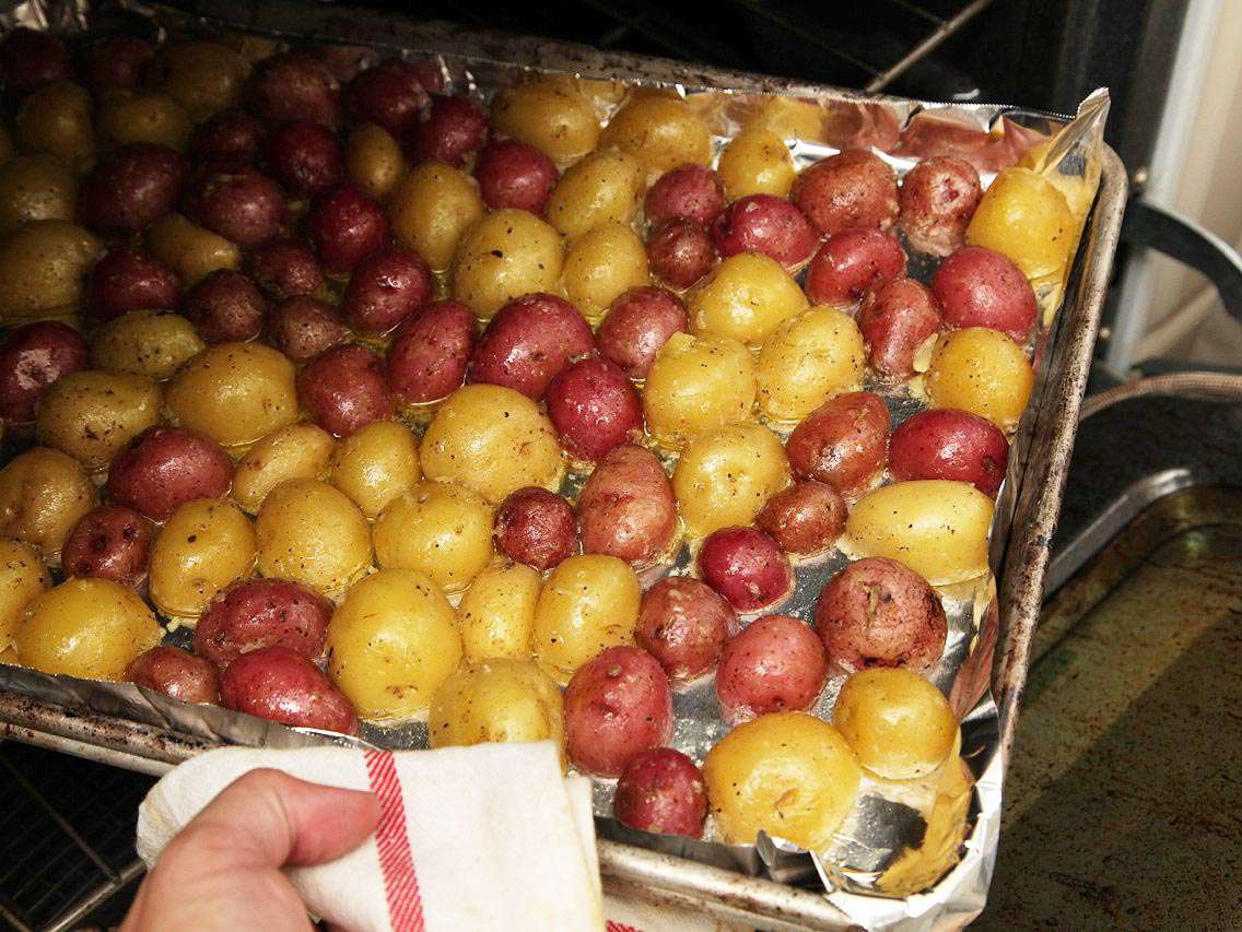 Hand holding baking sheet covered in red and yellow new potatoes sliding into oven