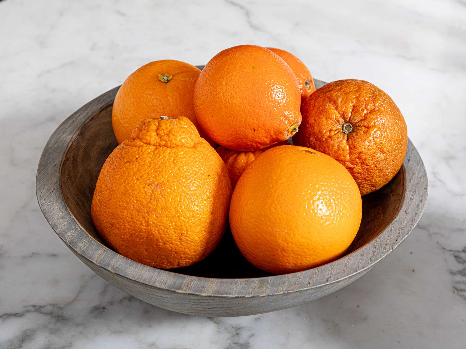 Oranges in a wooden bowl on top of a marble countertop.