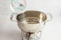 A person pouring water into a fondue pot's base using a measuring cup.