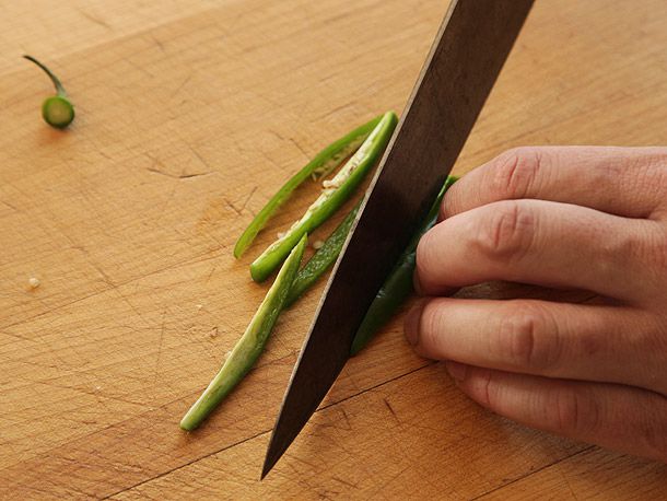 Tucked fingers against chef's knife cutting green pepper into thin strips on wooden surface