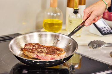 A hand holding a frying pan with a piece of steak being seared on a stovetop