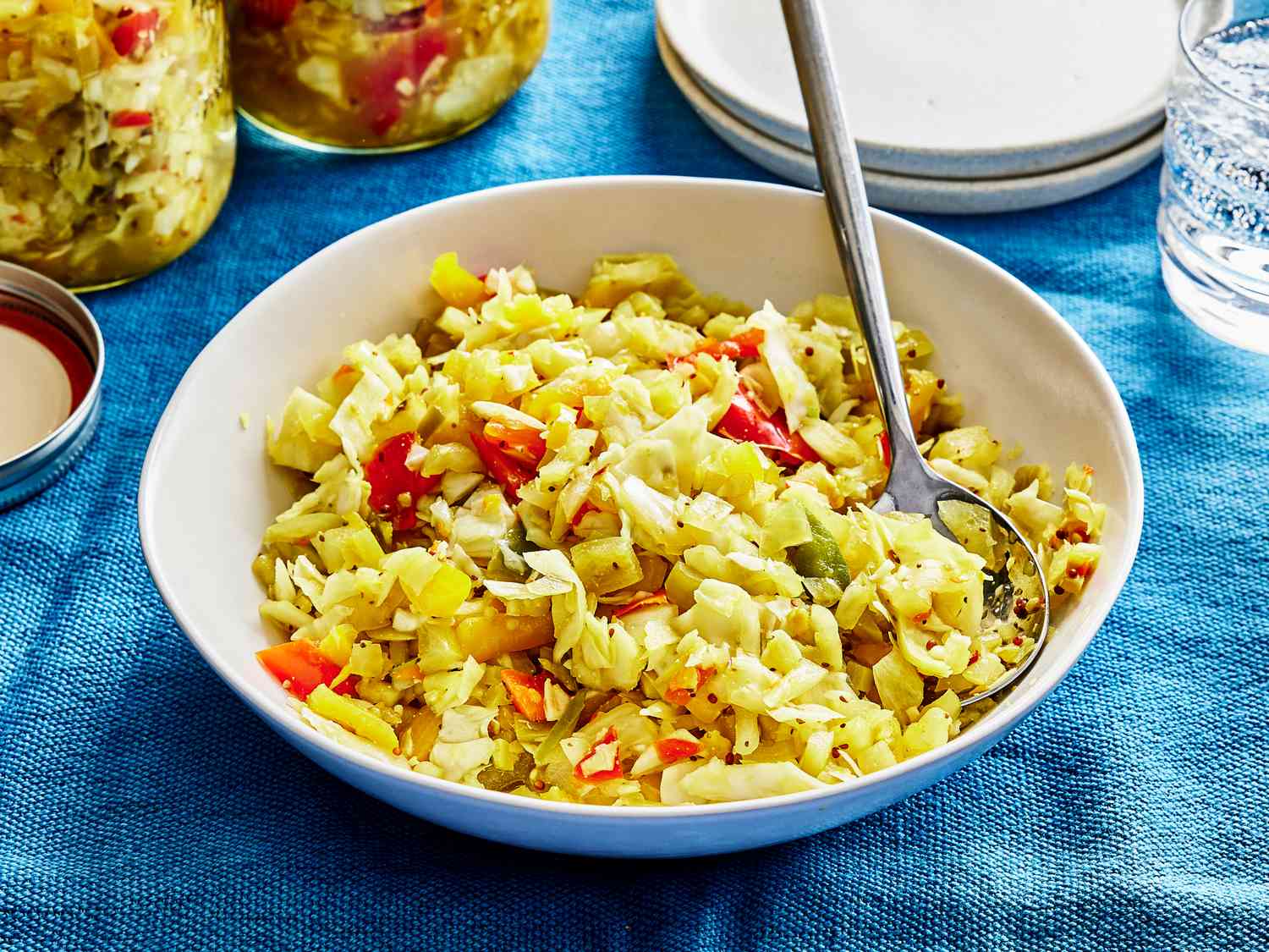 Bowl of Chow Chow with a spoon in it. The bowl is on a blue table cloth with other plates and sparkling water around it 