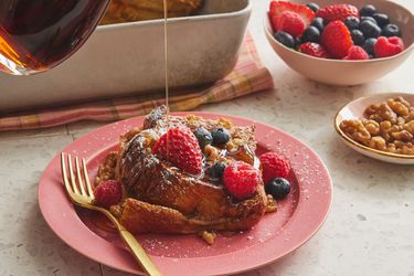 Gif of syrup pouring ontop of french toast casserole with berries, on a pink plate, with topping and casserole in the background