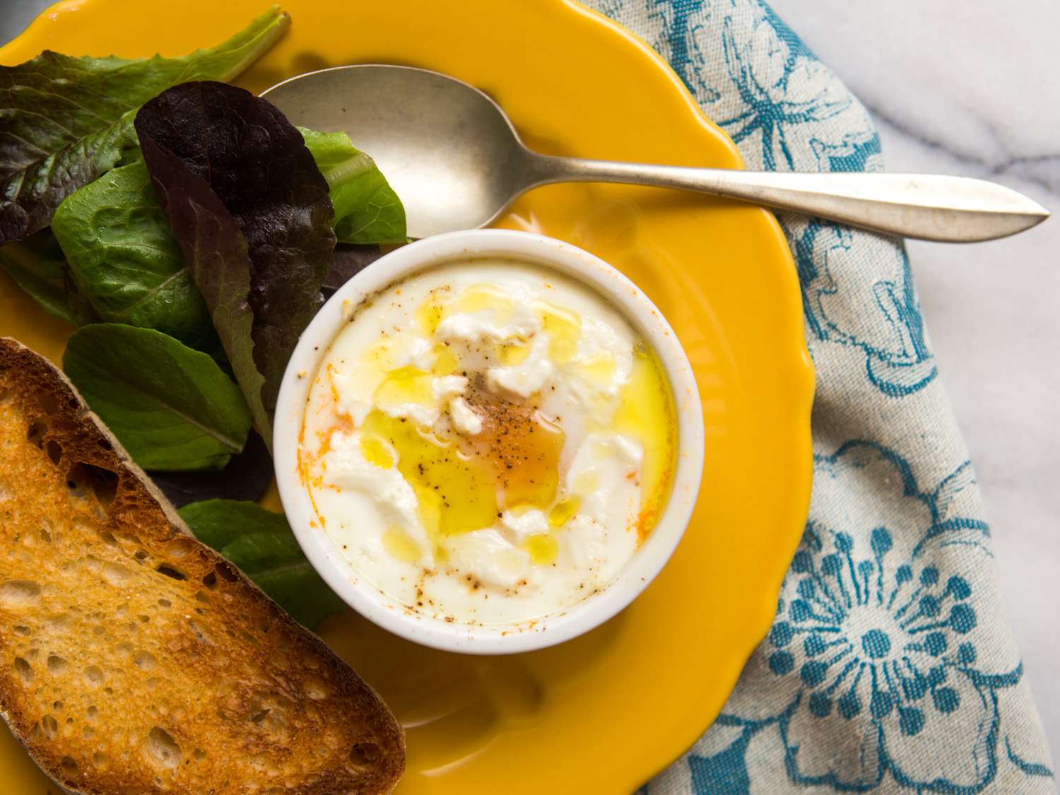 Overhead view of eggs en cocotte with tomato and goat cheese, served on a plate with a green salad and toast.
