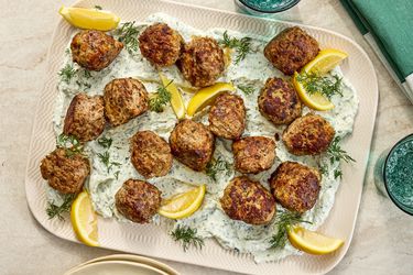 meatballs on top of yogurt with fresh herbs and lemon wedges on a platter. Side of sparkling water and green napkin on the side 