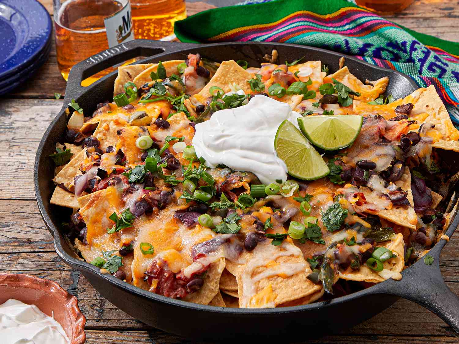 A skillet of nachos topped with melted cheese, black beans, sour cream, lime wedges, and green onions, placed on a wooden surface