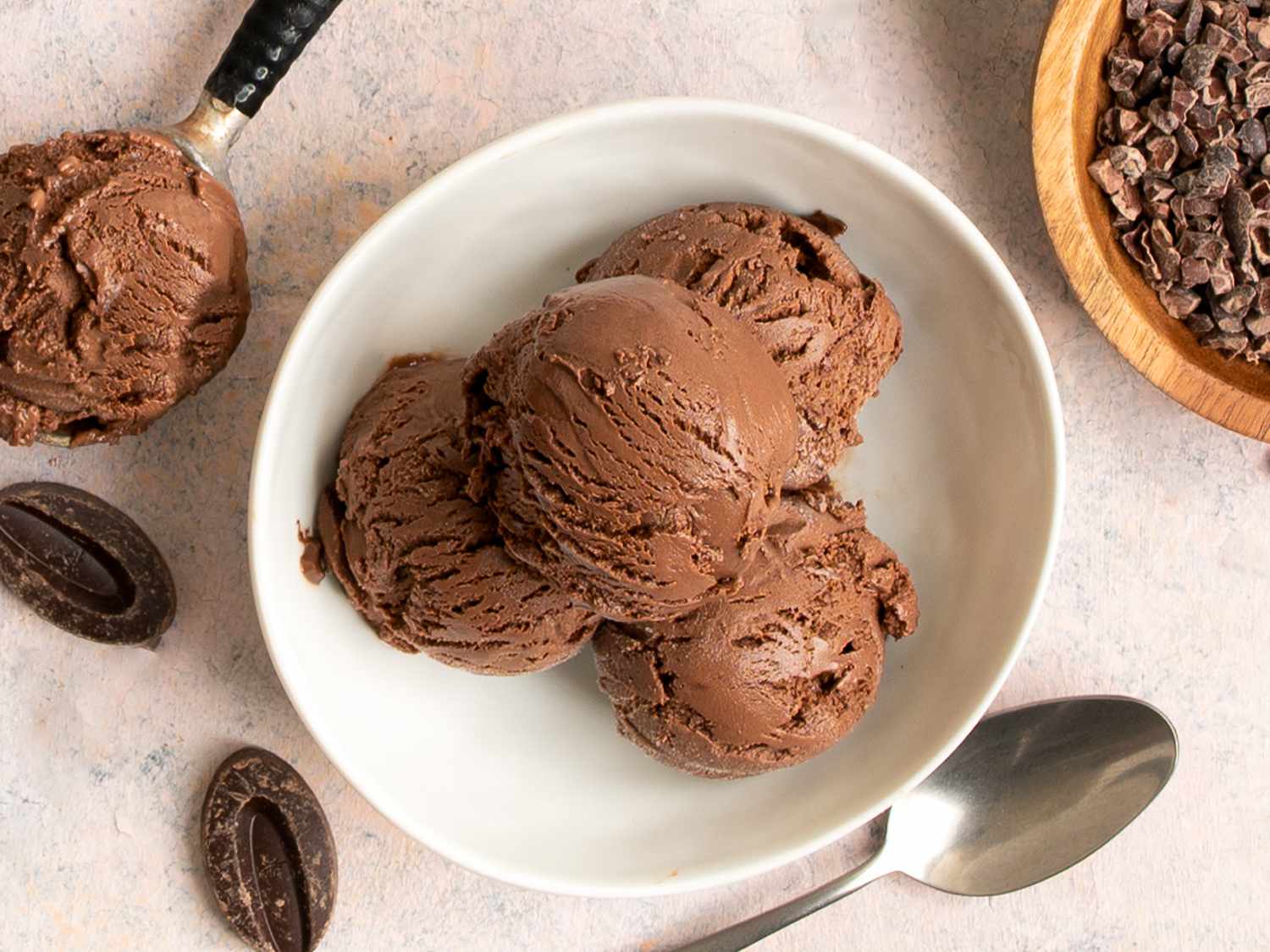 Four scoops of chocolate ice cream in a round white ceramic bowl. Around the periphery of the bowl are a metal spoon, a small wooden bowl holding cacao nibs, two pieces of chocolate, and a scoop holding ice cream.
