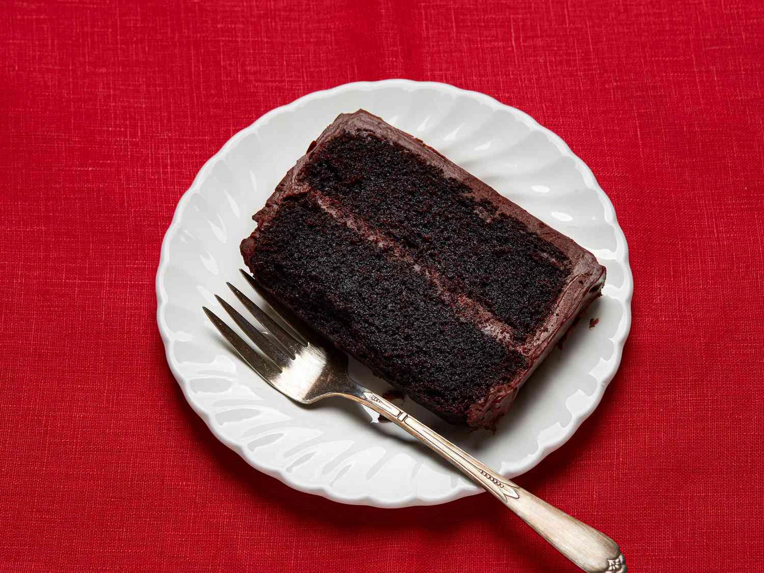 slice of chocolate cake on a white plate, with a fork, on a red fabric