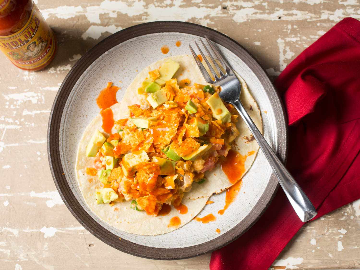 A plate of Doritos migas with hot sauce, a fork, and a red cloth napkin.