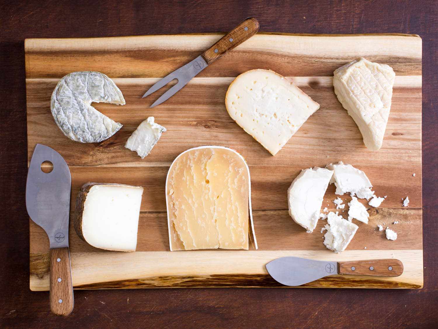 A group of six different cheeses on a cutting board with three cheese knives. 