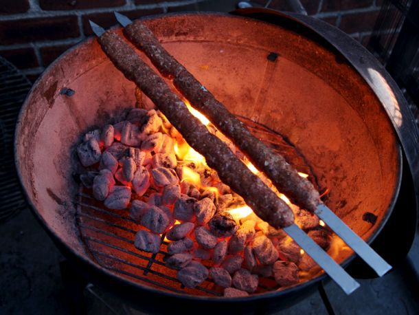 Adana kebabs being grilled over charcoal. The wide, flat metal skewers are long enough to set across the entire kettle, making a grill grate unnecessary.