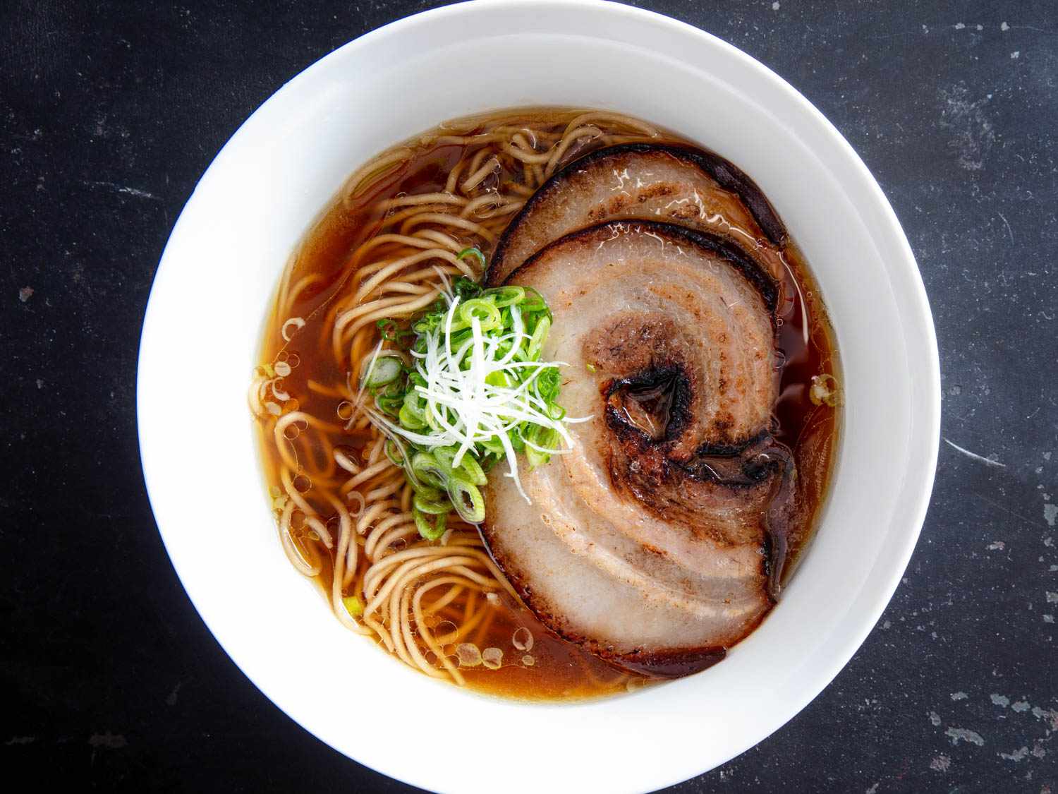 Overhead shot of a bowl of ramen in broth topped with sliced of pressure-cooked pork belly chashu and garnished with sliced scallion. 