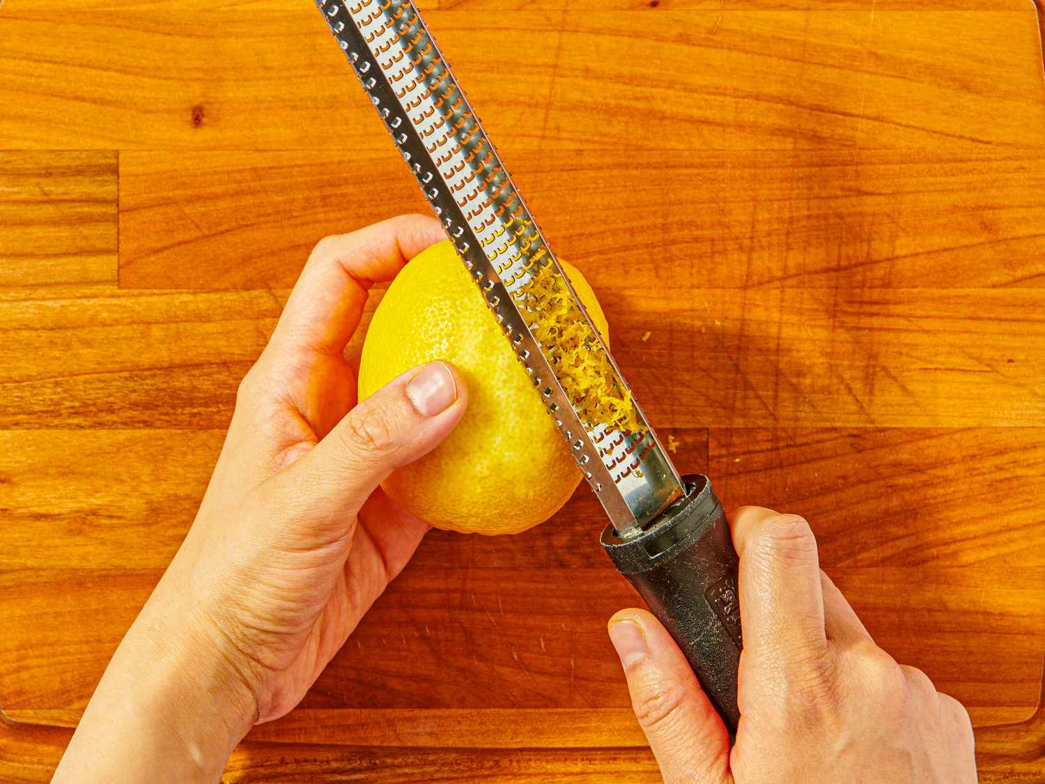 Hands grating the peel of a lemon with a zester on a wooden surface