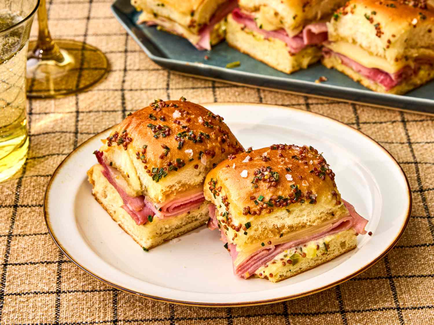 Ham and cheese sliders on a plate garnished with a poppy seed topping with additional sliders on a tray in the background