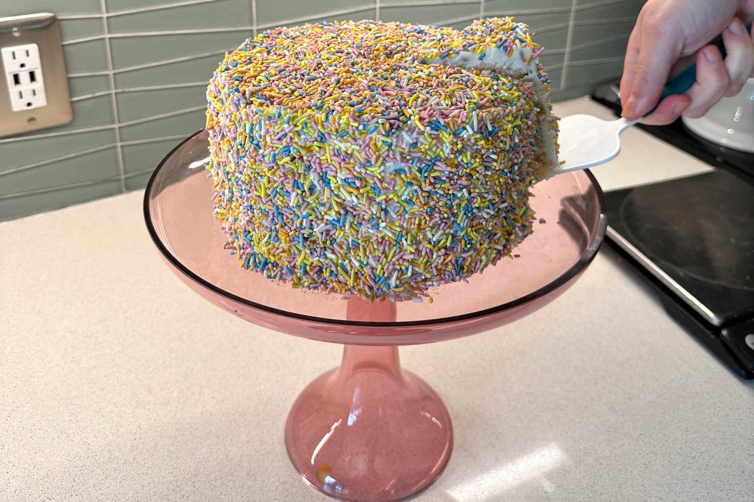 A cake, covered in sprinkles, sits on a pink glass cake stand