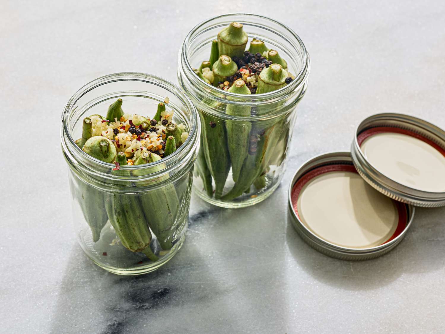 Two jars of pickled okra with seasonings on a surface lids placed aside