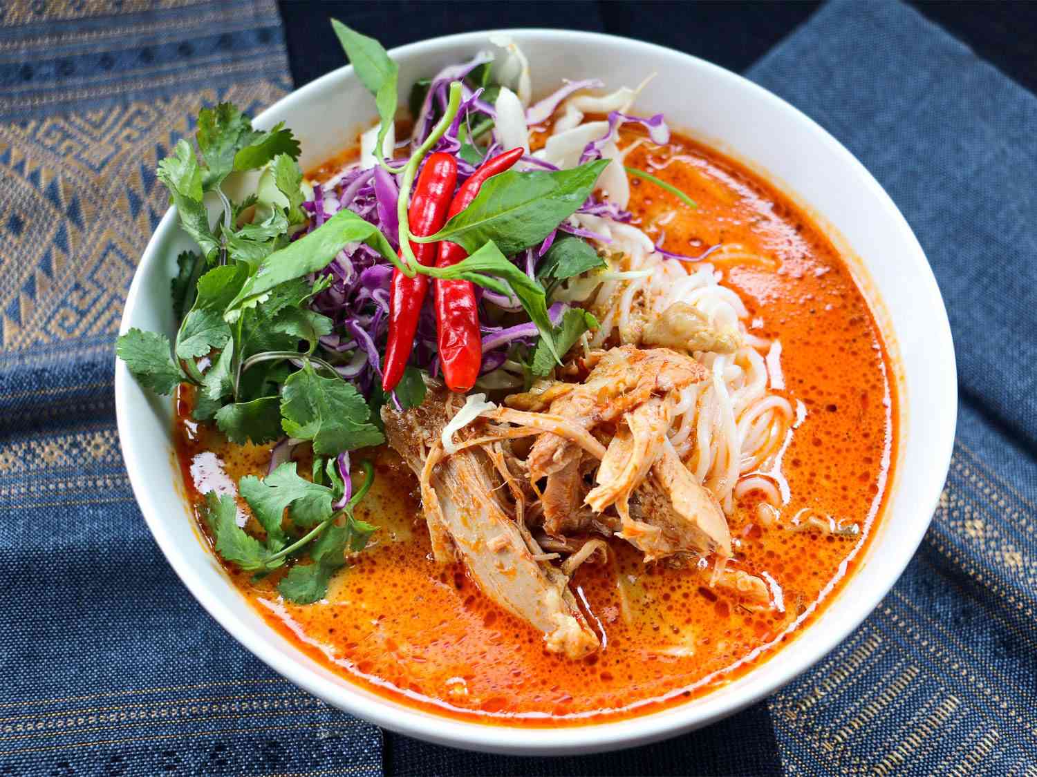 A bowl of khao poon: fermented vermicelli noodles in a bright red curry broth, with meat, herbs, red chilies, and purple cabbage.
