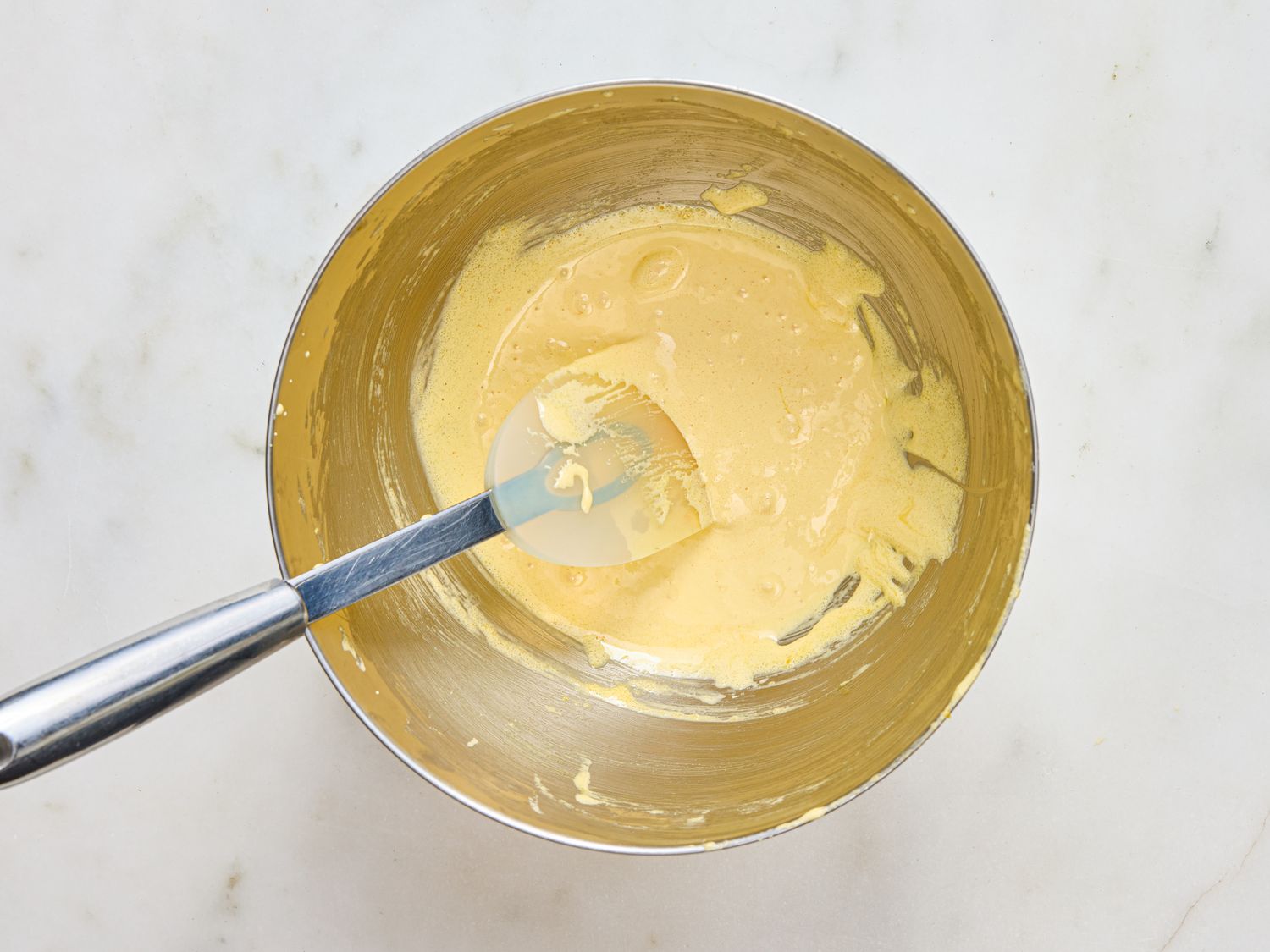 A metal mixing bowl containing a creamy mixture being stirred with a spatula