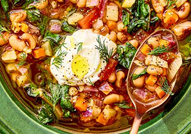 A bowl of stew with blackeyed peas vegetables greens and a dollop of cream topped with dill alongside a spoon