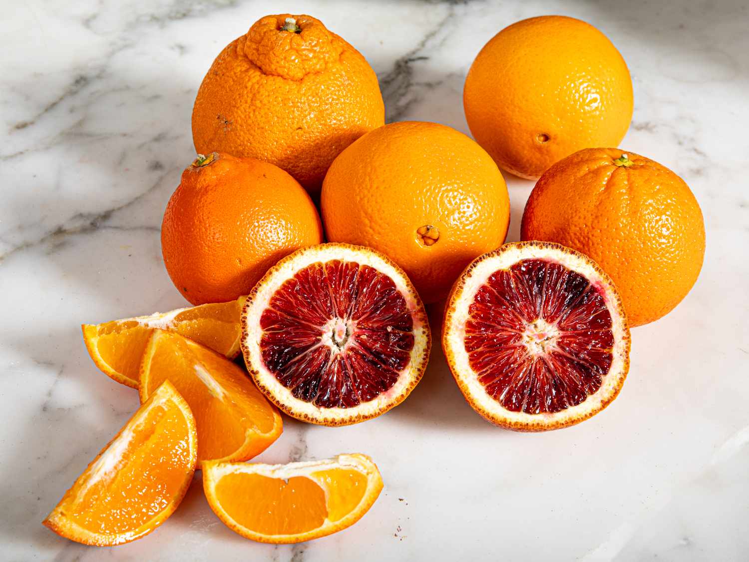 Whole and cut up oranges against a white marble countertop.