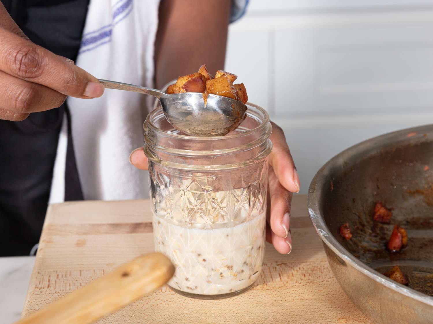 A person spooning spiced apples into a jar of overnight oats on a wooden surface