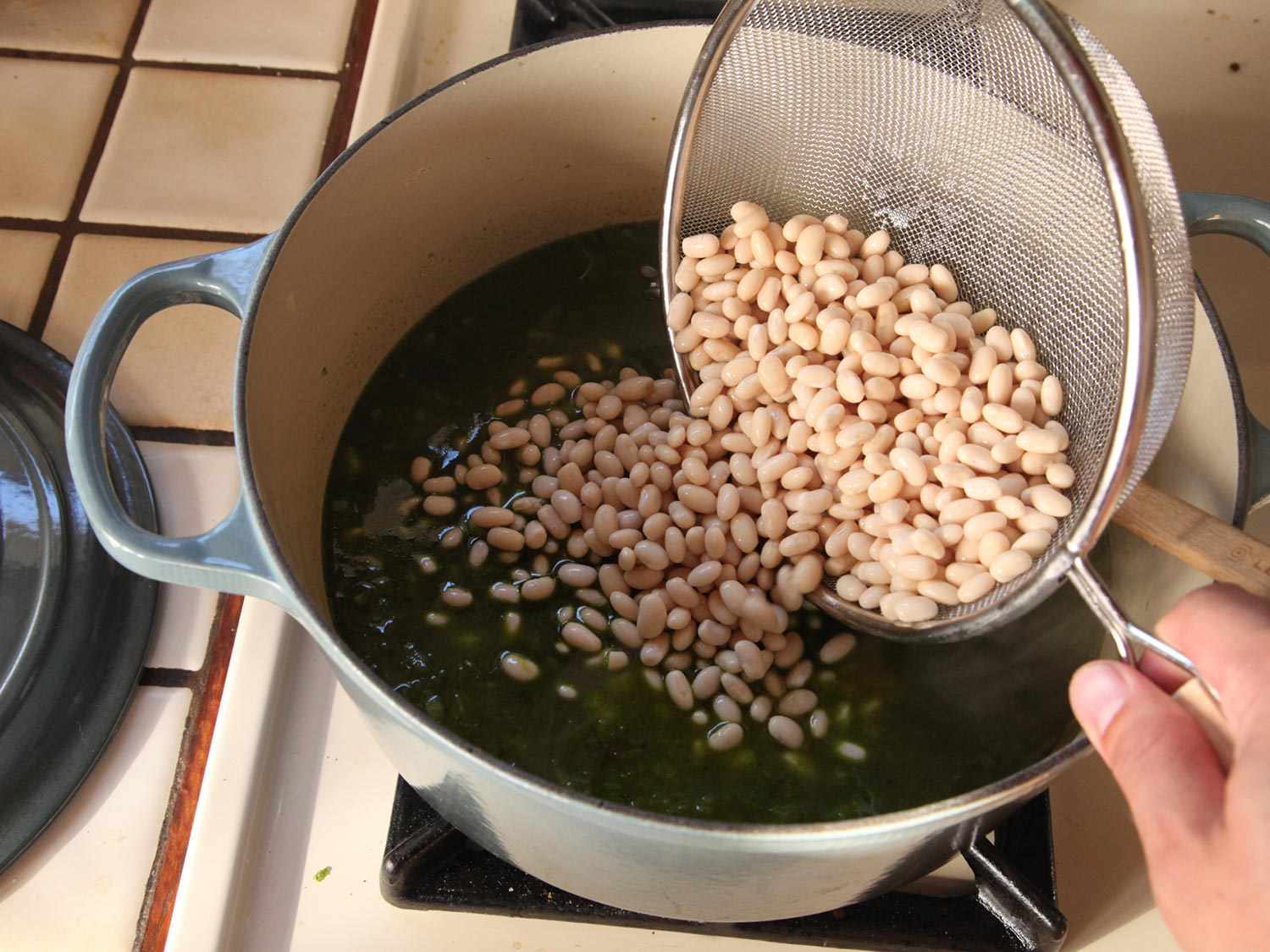 Transferring drained white beans into the pot of green chile base with a fine-mesh strainer
