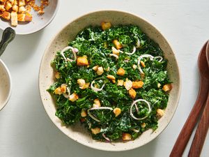 Kale caesar salad in a speckled ceramic bowl. On the left periphery are a pair of wooden serving spoons, and on the left periphery is a small bowl holding croutons.