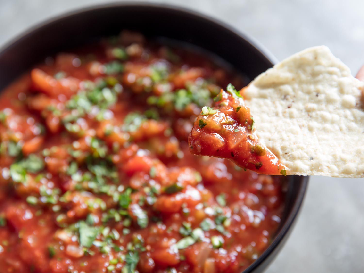 Close-up of a chip topped with salsa. A bowl is in the background, slightly out of focus.