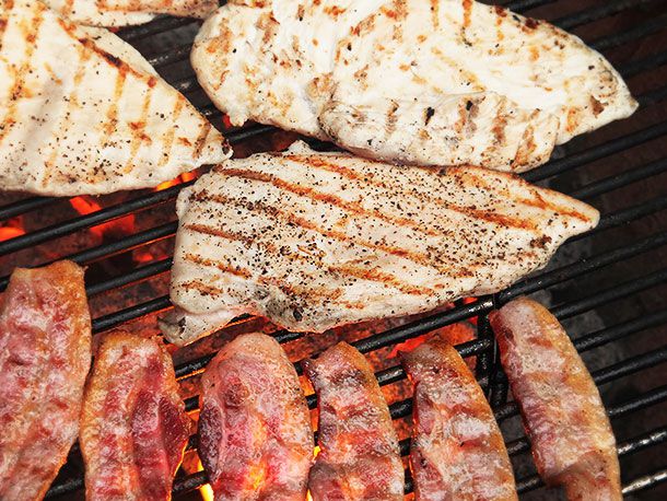 Chicken breast getting lightly charred over hot coals. Slices of bacon are off to one side of the coals getting browned and crispy.