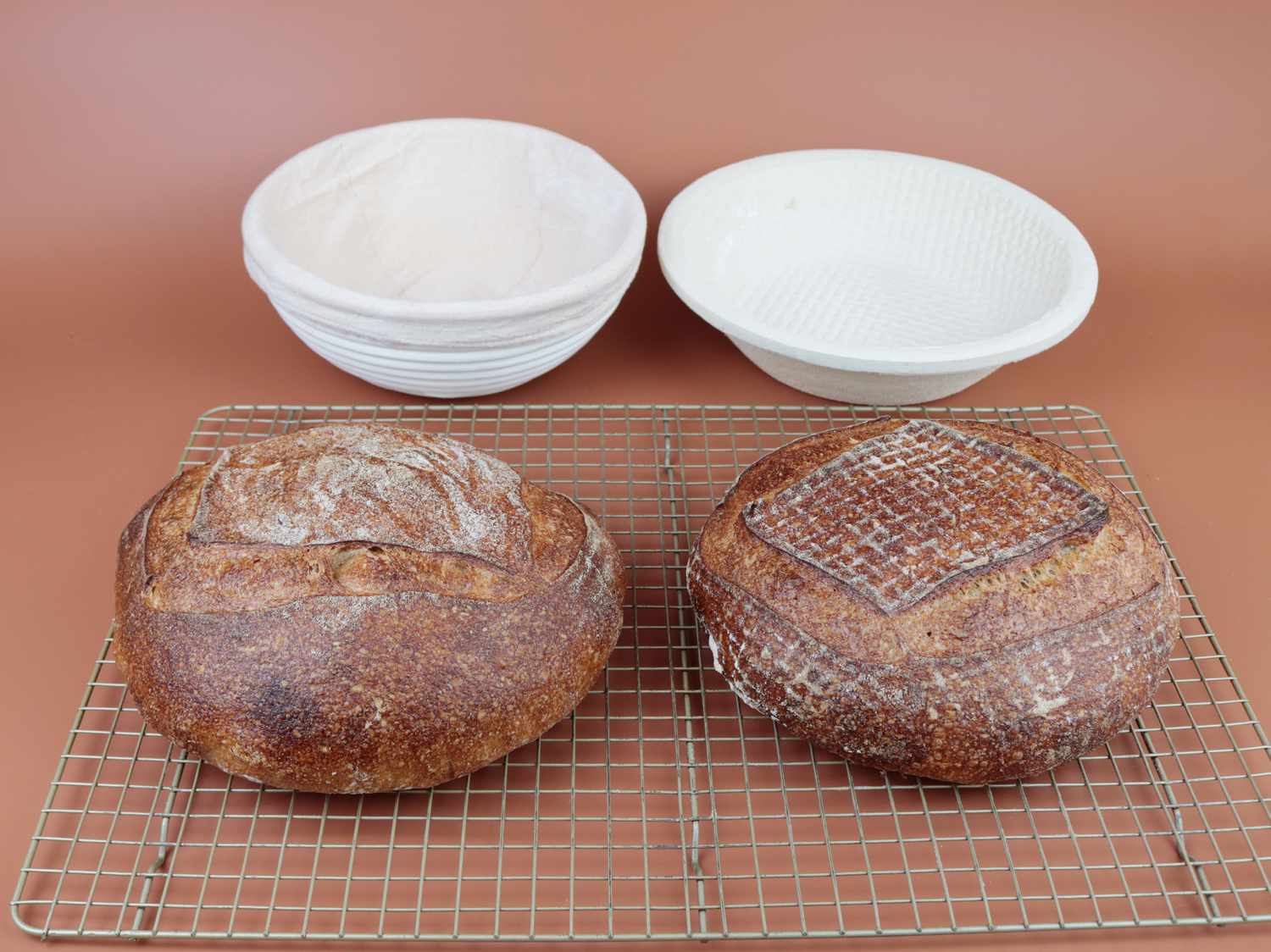 Two baked round loaves of bread on a cooling rack in front of two proofing baskets