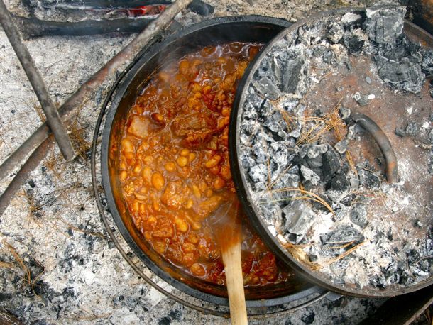 A Dutch oven filled with chili cooking over campfire coals and being stirred with a wooden spoon.