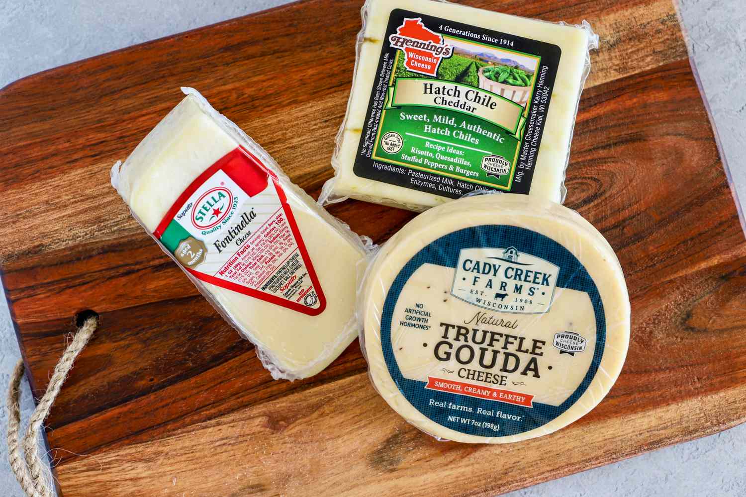A wooden board holding three types of packaged cheese from Wisconsin Cheese Mart's Wisconsin Cheese of the Month Club, including Fontinella, Hatch Chile Cheddar and Truffle Gouda
