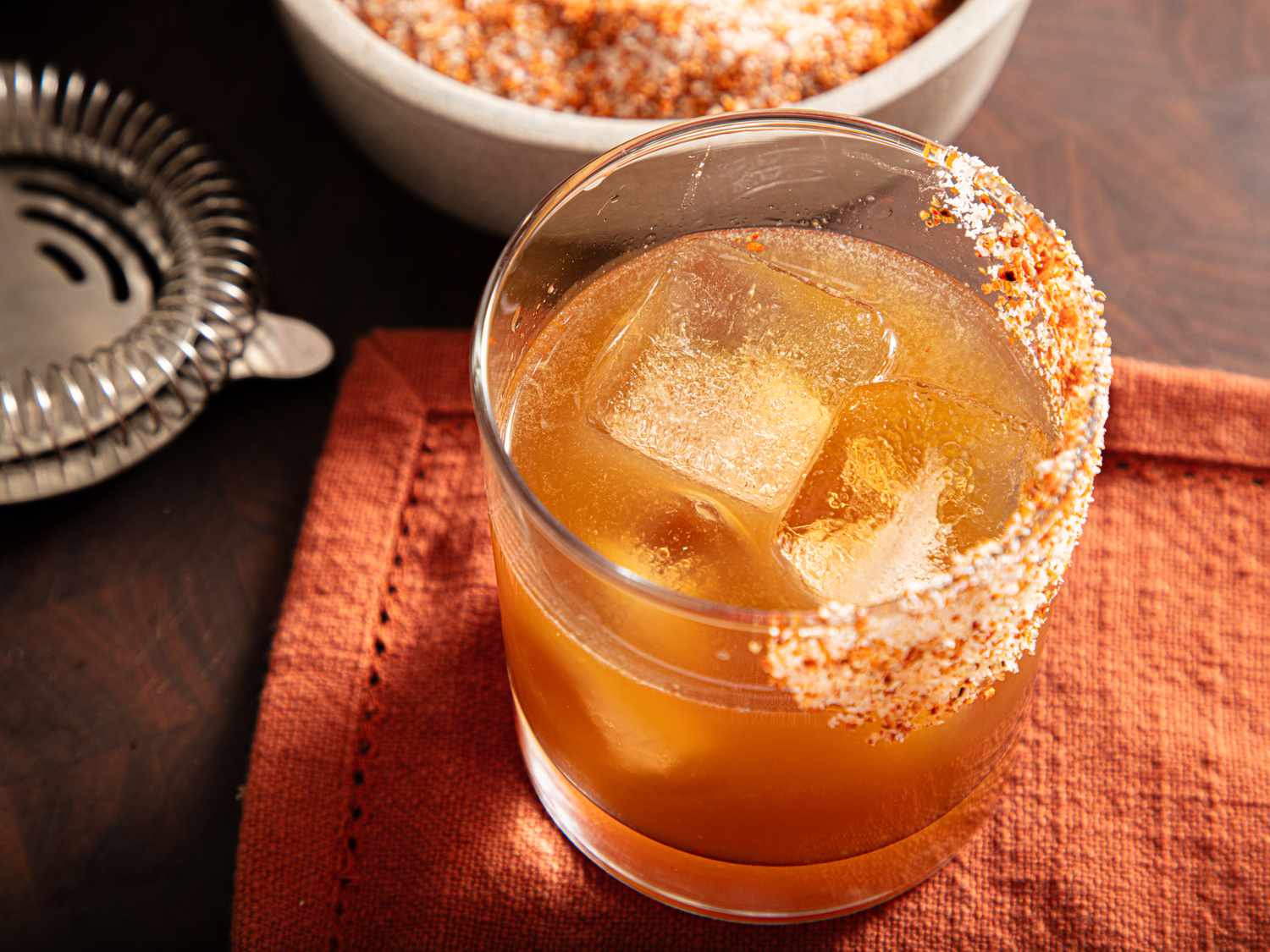 A cocktail glass filled with a beverage served over ice, rimmed with a spicy or seasoned salt, on an orange cloth