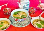A large bowl of longevity noodles are served on a red table, alongside a pot of tea. Diners serve themselves from the bowl, transferring the long noodles with chopsticks.