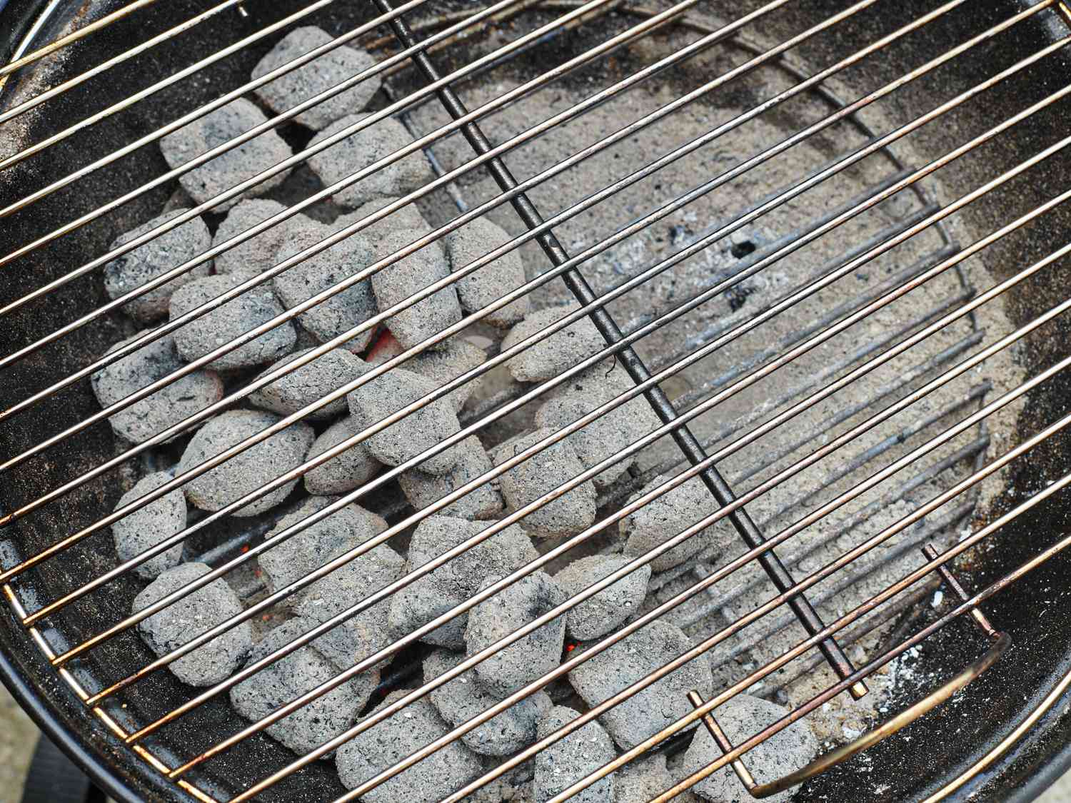 A round black charcoal grill, showing the ash-gray coals on the left side of the bottom grate, creating one zone for direct heat and one for indirect heat.