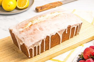 Lemon pound cake with glaze on a wooden board, surrounded by lemons, a knife, and a bowl of fresh berries.
