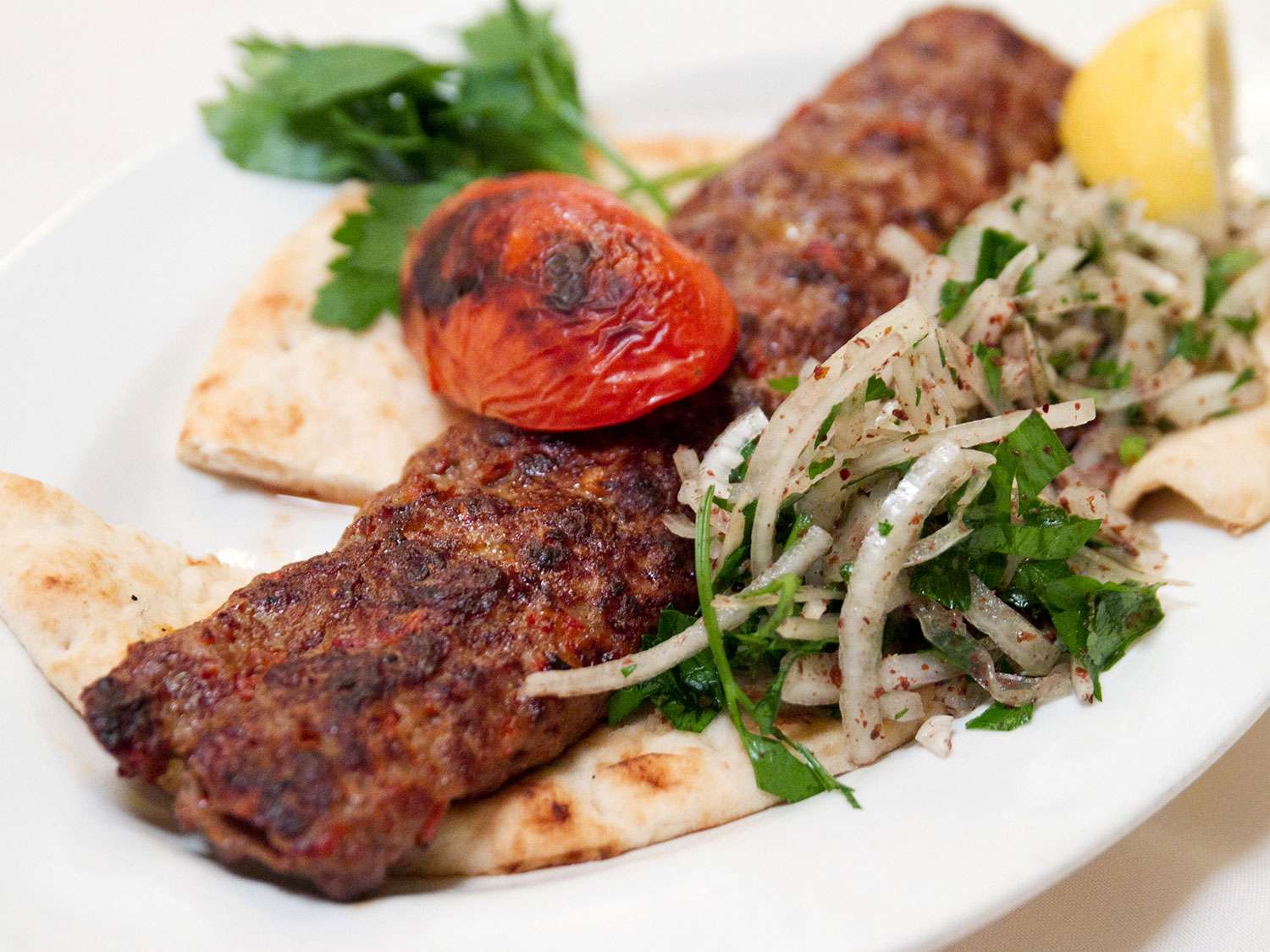 An adana kebab on a platter with pita bread, a grilled tomato, a lemon wedge, and an onion-parsley-sumac salad 