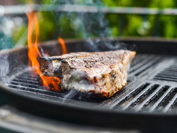 A porterhouse is being seared on the grill and flames are leaping from the bone side as some of the fat catches from the intense heat.