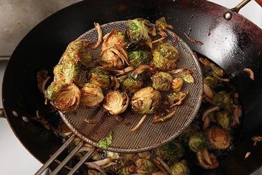 Halved Brussels sprouts and sliced shallots being deep-fried in a wok. A metal spider is holding some of the vegetables above the wok. 