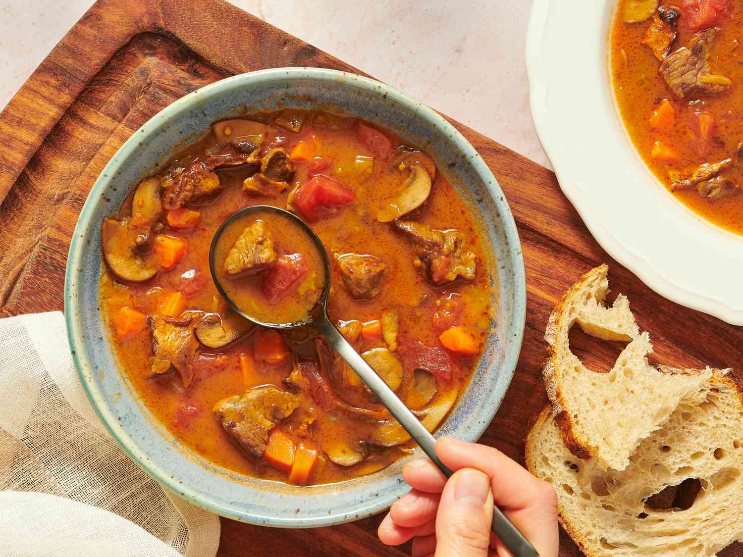 Kansas City Steak soup in bowl on wooden board wit ripped bread, with hand holding spoonful of soup