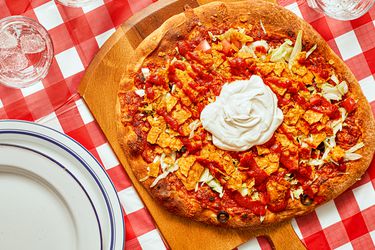 Taco pizza with sour cream on a wooden board, set on a red-checkered tablecloth, plates, and glasses nearby.