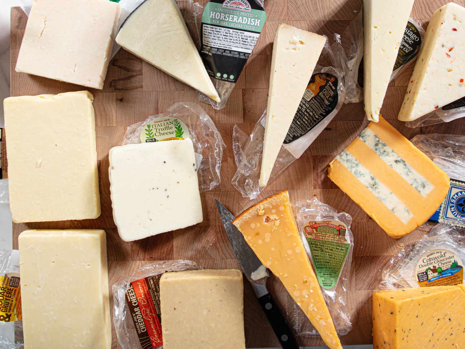 Various types of cheese displayed on a wooden surface with some packaging visible and a knife placed among the items
