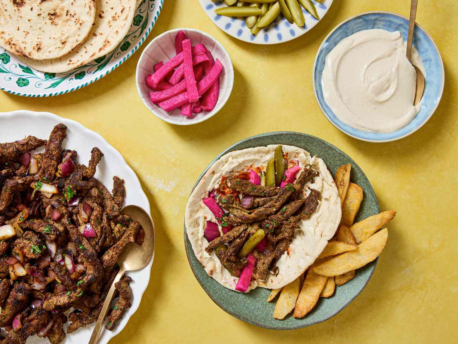 Beef shawarma wrap with accompaniments including pickles tahini sauce and pita bread on a table