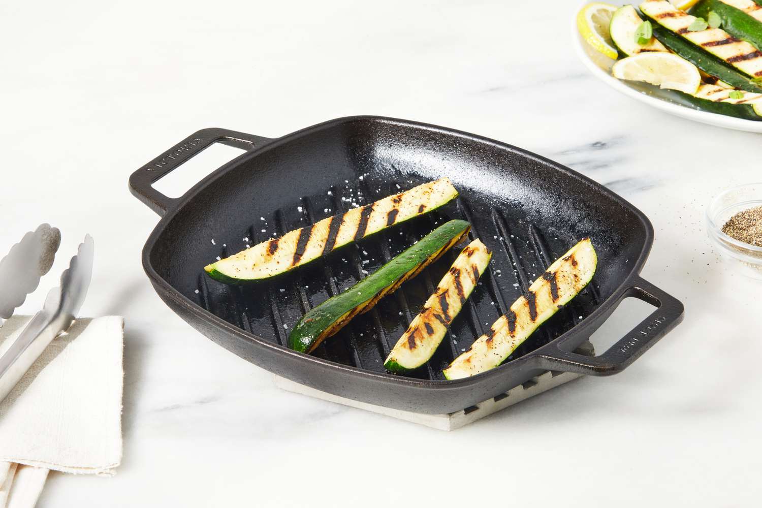The Victoria Cast Iron Square Grill Pan on a table with grilled zucchini inside the pan. 