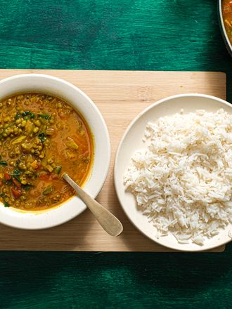 Bowl of Moong dal in a bowl, with a bowl of white rice next to it. Bowls are on a wooden board on top of green textured surface, and larger bowl of Dal is in the side of the frame. 