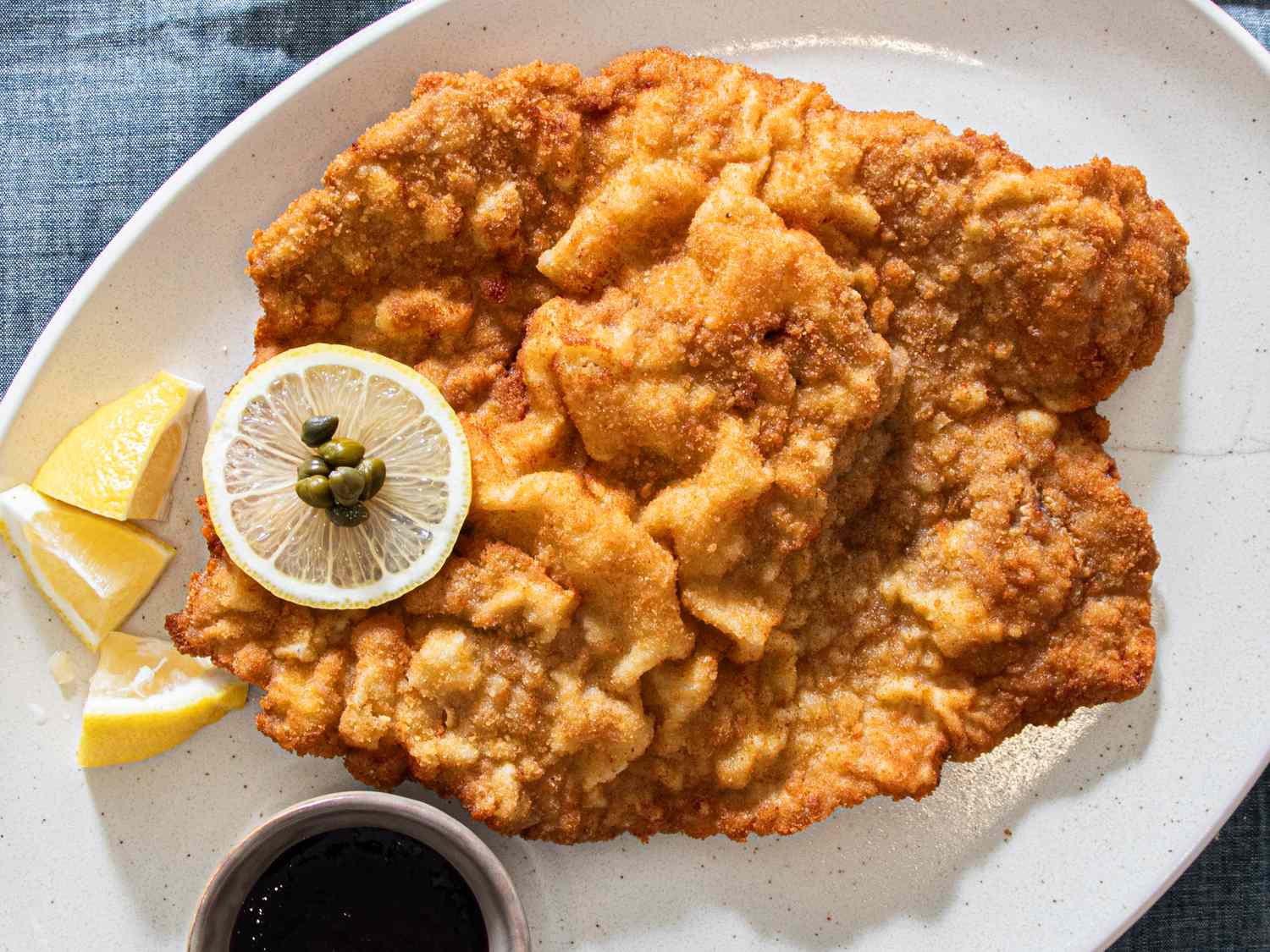 A plate of Wiener Schnitzel with a lemon slice and capers garnish accompanied by a cooked sauce served in a small dish