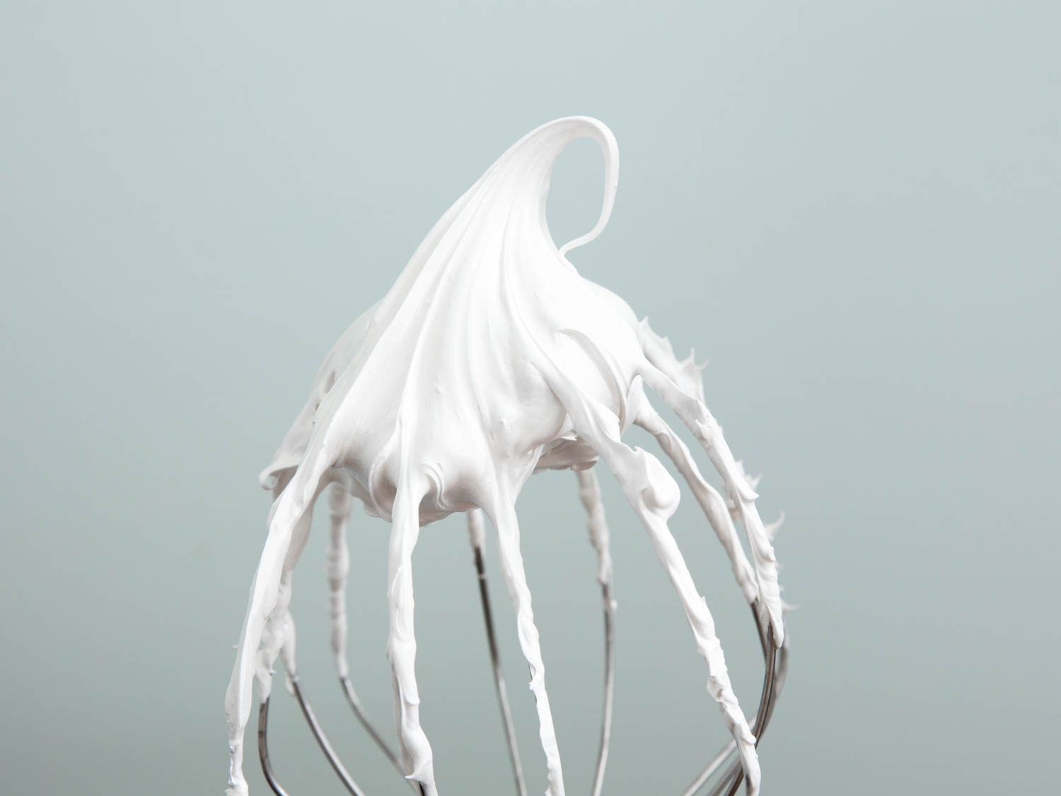 The wire whisk of stand mixer, with a glob of meringue caught in its wires, and a single curl of stiffly whipped meringue tilting to one side, like a curlicue of soft serve