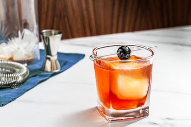 A Boulevardier cocktail in a cocktail glass with one large cube of ice, garnished with a cherry on a cocktail pick.