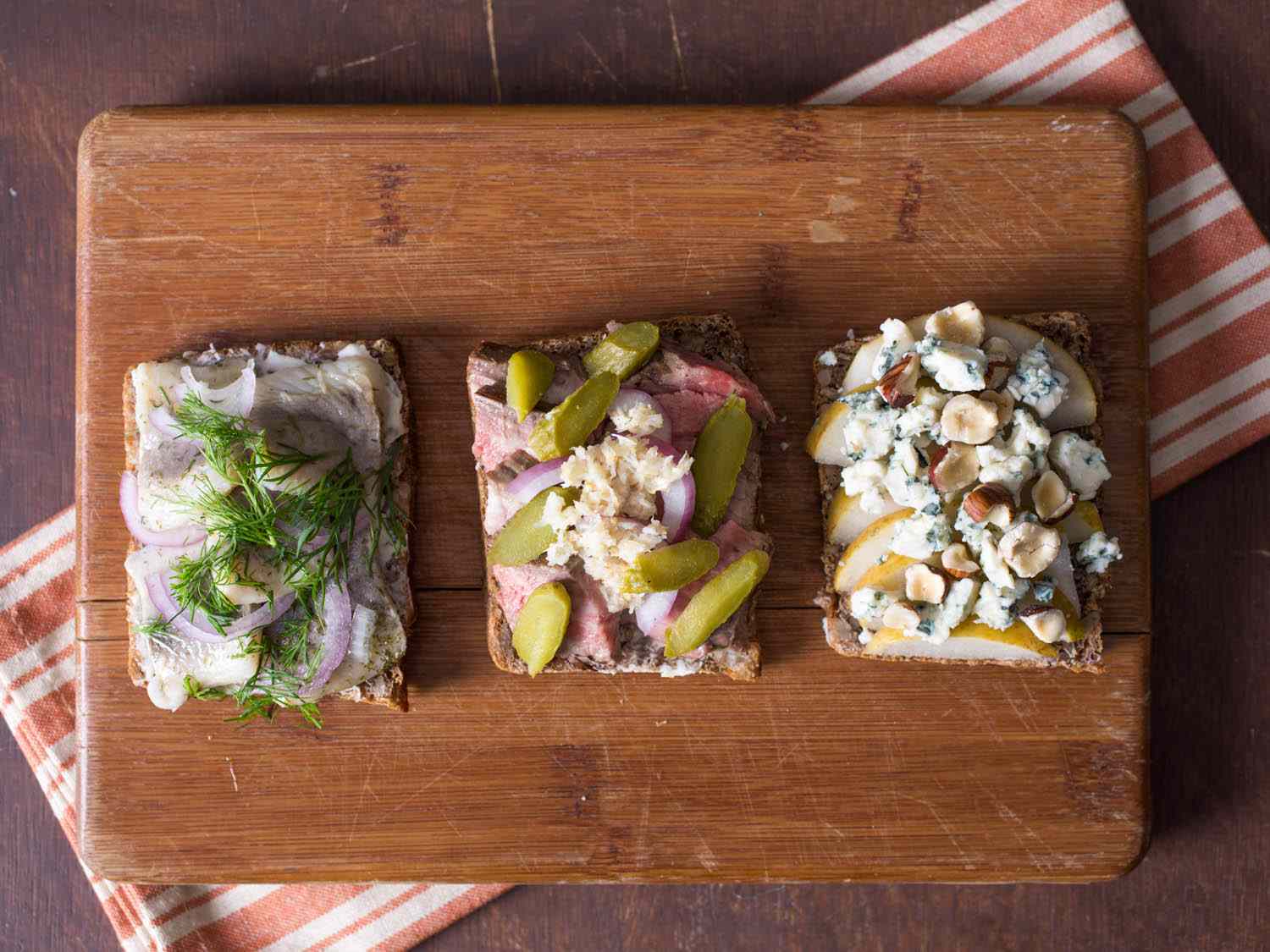 Three different Smørrebrød on a wood cutting board, each with different toppings.